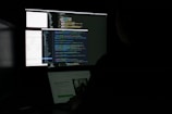 A person is sitting in front of two monitors displaying a dark-themed code editor with colorful lines of code. The setting is dimly lit, suggesting a focus on the screen's content.