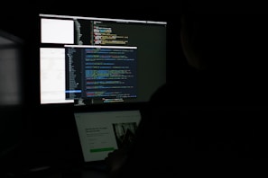 A person is sitting in front of two monitors displaying a dark-themed code editor with colorful lines of code. The setting is dimly lit, suggesting a focus on the screen's content.