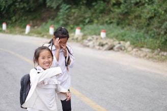Two children are walking along a rural roadside, both smiling and appearing to enjoy each other's company. They are wearing backpacks and casual clothing. The background features greenery and roadside markers.