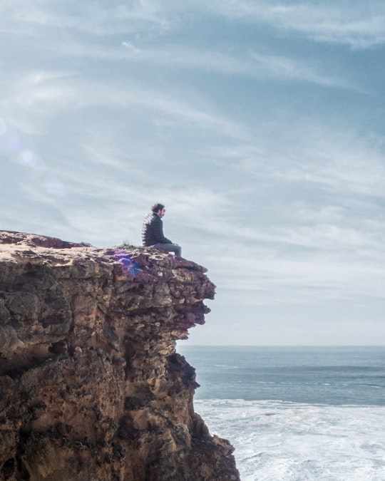 man in blue shirt sitting on brown rock formation during daytime in Nazaré Portugal
