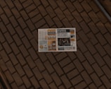 A newspaper lies open on a brick pavement. The paper appears to have a variety of articles, images, and advertisements, with the title 'Metro' visible on the front page. The brick pattern provides a textured, earthy backdrop for the newspaper.