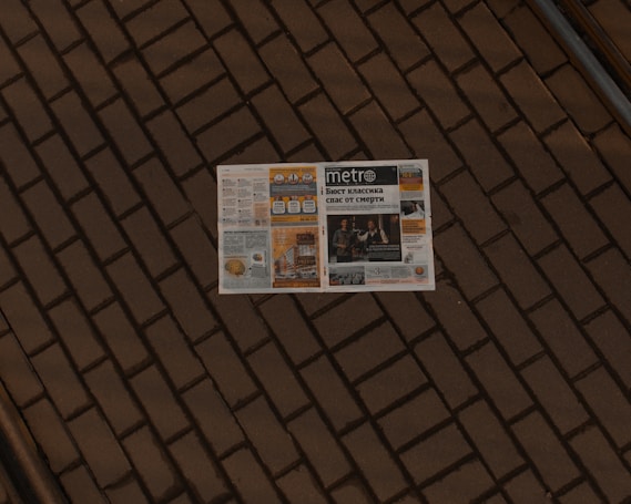 A newspaper lies open on a brick pavement. The paper appears to have a variety of articles, images, and advertisements, with the title 'Metro' visible on the front page. The brick pattern provides a textured, earthy backdrop for the newspaper.