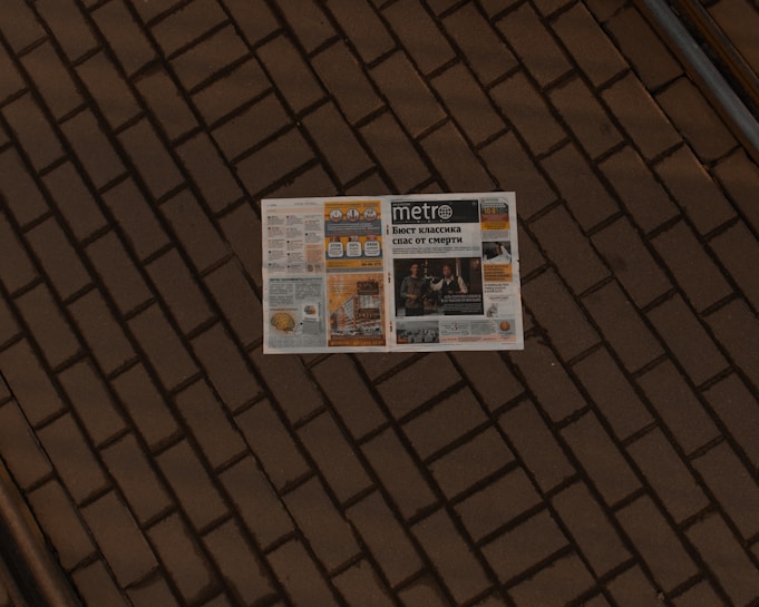 A newspaper lies open on a brick pavement. The paper appears to have a variety of articles, images, and advertisements, with the title 'Metro' visible on the front page. The brick pattern provides a textured, earthy backdrop for the newspaper.