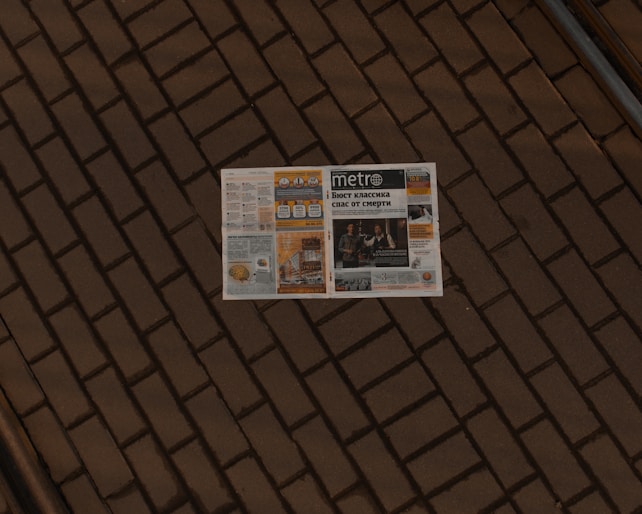 A newspaper lies open on a brick pavement. The paper appears to have a variety of articles, images, and advertisements, with the title 'Metro' visible on the front page. The brick pattern provides a textured, earthy backdrop for the newspaper.
