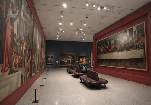A spacious art gallery featuring large classical paintings adorning the walls. Several benches are placed in the middle for visitors to sit and admire the artwork. The lighting is soft and focused, enhancing the details and colors of the paintings.