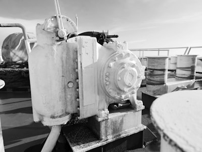 A black and white image featuring industrial machinery on what appears to be a ship deck. Central to the image is a large, cylindrical mechanical device with various knobs, bolts, and piping. Surrounding the machinery are ropes and other mechanical components. The background reveals parts of the ship's railings and a clear sky.