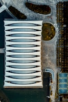An aerial view of a modern architectural building with a unique, wavy roof design surrounded by an urban landscape. The structure is adjacent to a body of water, with landscaped areas and a series of basketball courts visible nearby. The area features various geometric patterns and sections of greenery.