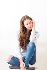A young person with long curly brown hair and wearing a striped long-sleeve shirt and ripped blue jeans. They are sitting cross-legged on a cushioned stool, looking pensively to the side with one hand resting on their cheek.