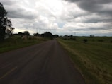 A rural road extends into the distance, surrounded by grassy fields and trees. The sky is overcast with dark clouds, suggesting an impending storm. Small buildings and utility poles are visible along the road.
