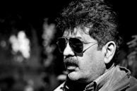 A man with curly hair and a mustache is wearing dark aviator sunglasses and a collared shirt. The image is in black and white, with a strong contrast between light and shadow, giving dramatic emphasis on facial features.