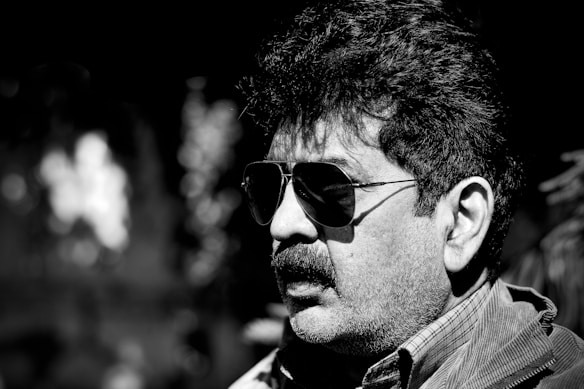 A man with curly hair and a mustache is wearing dark aviator sunglasses and a collared shirt. The image is in black and white, with a strong contrast between light and shadow, giving dramatic emphasis on facial features.