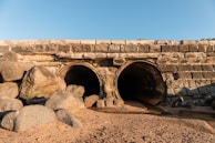 Two large stone drainage tunnels are situated beneath a stone bridge, with a sandy shore and scattered rocks leading up to the openings. The scene is set under a clear blue sky.