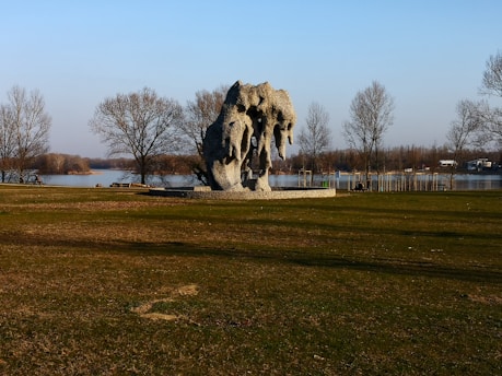 A large, abstract sculpture made of stone or concrete stands in an open grassy area, surrounded by leafless trees. The sculpture has irregular, draped forms, creating a dynamic silhouette against a clear sky. In the background, a river or lake can be seen, and there are sparse trees lining its banks.
