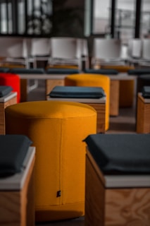 A modern interior setting featuring rows of colorful ottomans, primarily in bright yellow and red, arranged beside wooden tables with dark cushions on top. The soft lighting and arrangement suggest a conference or lecture setup, with white chairs visible in the background.