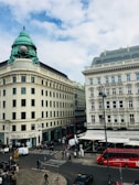 A bustling city intersection with historic buildings, featuring a corner tourist information office and a cafe with outdoor seating. A red double-decker tourist bus is parked nearby, and people are walking across the street. The architecture includes ornate facades and a distinctive copper dome atop one building against a backdrop of a partly cloudy sky.