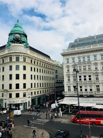 A bustling city intersection with historic buildings, featuring a corner tourist information office and a cafe with outdoor seating. A red double-decker tourist bus is parked nearby, and people are walking across the street. The architecture includes ornate facades and a distinctive copper dome atop one building against a backdrop of a partly cloudy sky.