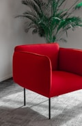 A red armchair with sleek, minimalist design stands on a textured gray carpet. Behind the chair, a tall green indoor plant adds a touch of nature to the scene, set against a neutral-toned wall.