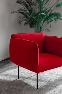A red armchair with sleek, minimalist design stands on a textured gray carpet. Behind the chair, a tall green indoor plant adds a touch of nature to the scene, set against a neutral-toned wall.
