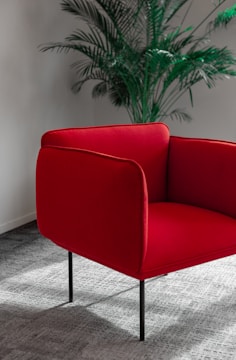 A red armchair with sleek, minimalist design stands on a textured gray carpet. Behind the chair, a tall green indoor plant adds a touch of nature to the scene, set against a neutral-toned wall.