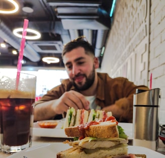 A person sits at a table in a cafe with food in the foreground including a sandwich and a glass of iced coffee. The cafe interior features circular ceiling lights and has a modern design.