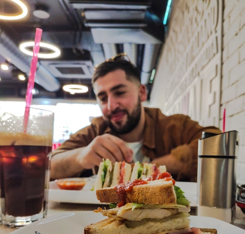A person sits at a table in a cafe with food in the foreground including a sandwich and a glass of iced coffee. The cafe interior features circular ceiling lights and has a modern design.