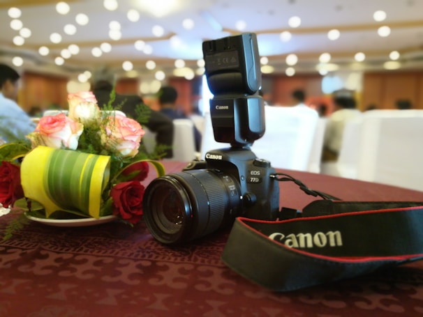 A Canon EOS 77D camera with a flash attachment rests on a maroon tablecloth. Nearby, a small bouquet of white and red roses adds a touch of elegance. In the background, a softly focused gathering of people is visible, indicating a possible event or meeting.
