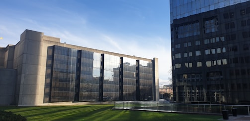 Two modern office buildings with reflective glass facades are positioned on either side of a well-maintained lawn. The building on the left is a monolithic concrete structure with multiple glass panels, while the building on the right has a darker, sleek appearance. The sky is clear with a few light clouds.