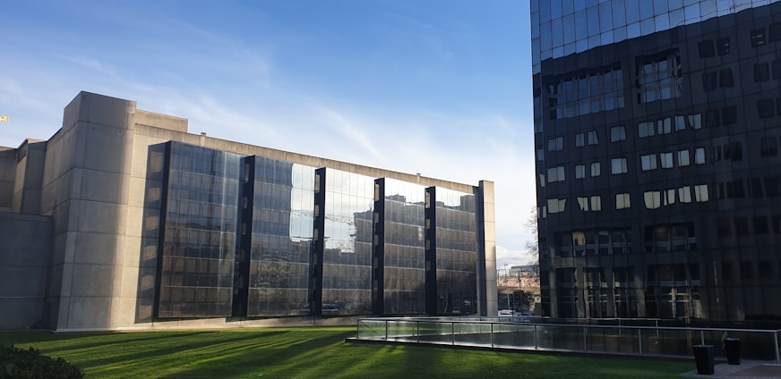 Two modern office buildings with reflective glass facades are positioned on either side of a well-maintained lawn. The building on the left is a monolithic concrete structure with multiple glass panels, while the building on the right has a darker, sleek appearance. The sky is clear with a few light clouds.