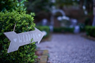 A wooden sign with the word 'wedding' and hearts points to a direction, attached to a lush green bush. The path beside it is gravel, leading to an area that appears to be decorated for an outdoor event, perhaps a wedding.