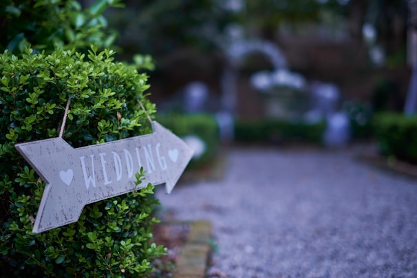 A wooden sign with the word 'wedding' and hearts points to a direction, attached to a lush green bush. The path beside it is gravel, leading to an area that appears to be decorated for an outdoor event, perhaps a wedding.