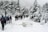 A group of people with backpacks and trekking poles are walking through a snowy forest path. Tall pine trees covered in snow line both sides of the path, creating a wintery landscape. The scene is set on an overcast day, with grey clouds adding to the wintry atmosphere.