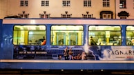 A blue tram is parked on a street with an advertisement for a Netflix series. The advertisement features a group of people, a stormy background with lightning, and text in another language. Inside the tram, passengers are visible sitting on the seats.