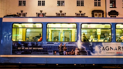 A blue tram is parked on a street with an advertisement for a Netflix series. The advertisement features a group of people, a stormy background with lightning, and text in another language. Inside the tram, passengers are visible sitting on the seats.