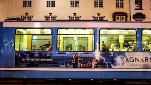 A blue tram is parked on a street with an advertisement for a Netflix series. The advertisement features a group of people, a stormy background with lightning, and text in another language. Inside the tram, passengers are visible sitting on the seats.