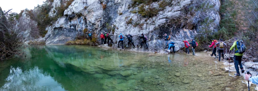 A group of hikers wearing backpacks and outdoor gear navigate a rocky path along the edge of a clear green body of water. They are surrounded by steep rock faces and sparse vegetation, indicating a rugged, natural environment.