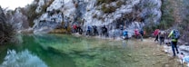 A group of hikers wearing backpacks and outdoor gear navigate a rocky path along the edge of a clear green body of water. They are surrounded by steep rock faces and sparse vegetation, indicating a rugged, natural environment.