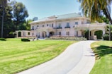 A large, elegant mansion with a two-story design is situated in a lush, well-maintained garden. The house features a light facade, green roof tiles, and an arched entrance with columns. A long, curved driveway approaches the house, and there are palm trees and trimmed hedges in the foreground.