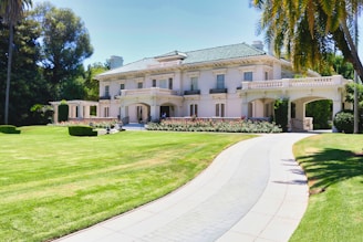 A large, elegant mansion with a two-story design is situated in a lush, well-maintained garden. The house features a light facade, green roof tiles, and an arched entrance with columns. A long, curved driveway approaches the house, and there are palm trees and trimmed hedges in the foreground.