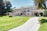 A large, elegant mansion with a two-story design is situated in a lush, well-maintained garden. The house features a light facade, green roof tiles, and an arched entrance with columns. A long, curved driveway approaches the house, and there are palm trees and trimmed hedges in the foreground.