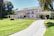 A large, elegant mansion with a two-story design is situated in a lush, well-maintained garden. The house features a light facade, green roof tiles, and an arched entrance with columns. A long, curved driveway approaches the house, and there are palm trees and trimmed hedges in the foreground.