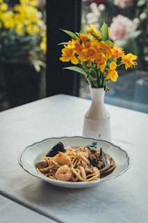 A bowl of seafood pasta is placed on a table adorned with a delicate arrangement of yellow flowers in a white vase. The dish includes shrimp, mussels, and pasta garnished with herbs, creating an appetizing appearance.