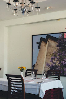 A dining table covered with a white tablecloth is set with cutlery, glasses, and a folded pink napkin. A small vase with yellow flowers decorates the center. The background features a large framed photograph of a building with lush purple foliage. Above, a modern chandelier adds elegance to the setting.