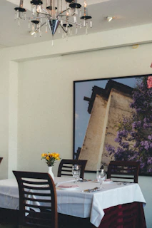 A dining table covered with a white tablecloth is set with cutlery, glasses, and a folded pink napkin. A small vase with yellow flowers decorates the center. The background features a large framed photograph of a building with lush purple foliage. Above, a modern chandelier adds elegance to the setting.