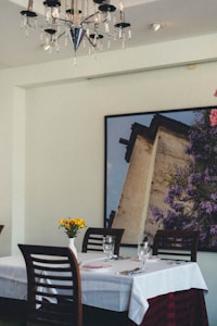 A dining table covered with a white tablecloth is set with cutlery, glasses, and a folded pink napkin. A small vase with yellow flowers decorates the center. The background features a large framed photograph of a building with lush purple foliage. Above, a modern chandelier adds elegance to the setting.