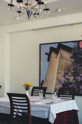 A dining table covered with a white tablecloth is set with cutlery, glasses, and a folded pink napkin. A small vase with yellow flowers decorates the center. The background features a large framed photograph of a building with lush purple foliage. Above, a modern chandelier adds elegance to the setting.