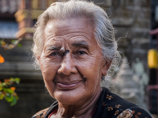 An elderly person with silver hair is smiling gently, conveying warmth and kindness. The person's face is framed by subtle lines that reflect a life of wisdom and experience. The background is softly blurred, hinting at an outdoor setting with foliage, adding a natural touch to the portrait.