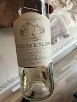 A wine bottle labeled 'Côtes de Bergerac Moelleux 2018' held in a hand. The label features a drawing of a chateau with trees and a fountain, and is marked 'Produit de France'. It's a clear glass bottle, giving a view of the contents inside.