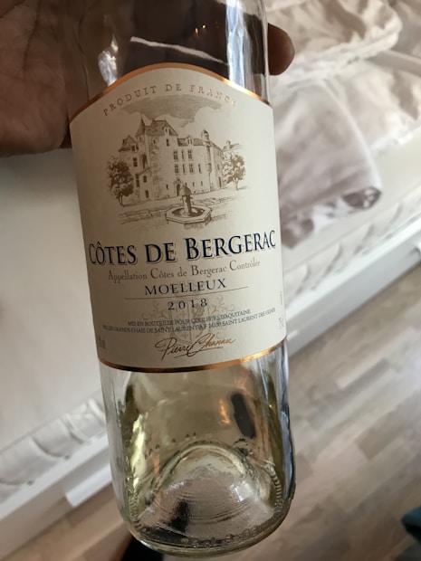 A wine bottle labeled 'C&ocirc;tes de Bergerac Moelleux 2018' held in a hand. The label features a drawing of a chateau with trees and a fountain, and is marked 'Produit de France'. It's a clear glass bottle, giving a view of the contents inside.