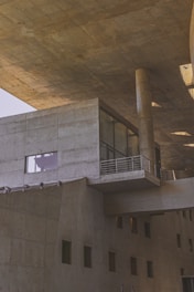 A modern architectural structure featuring large concrete surfaces, angular lines, and a robust design. The building includes various geometric shapes, such as rectangular windows and cylindrical columns, providing a sense of solidity and industrial aesthetic. The lighting casts subtle shadows on the textured concrete, enhancing the architectural details.