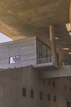 A modern architectural structure featuring large concrete surfaces, angular lines, and a robust design. The building includes various geometric shapes, such as rectangular windows and cylindrical columns, providing a sense of solidity and industrial aesthetic. The lighting casts subtle shadows on the textured concrete, enhancing the architectural details.